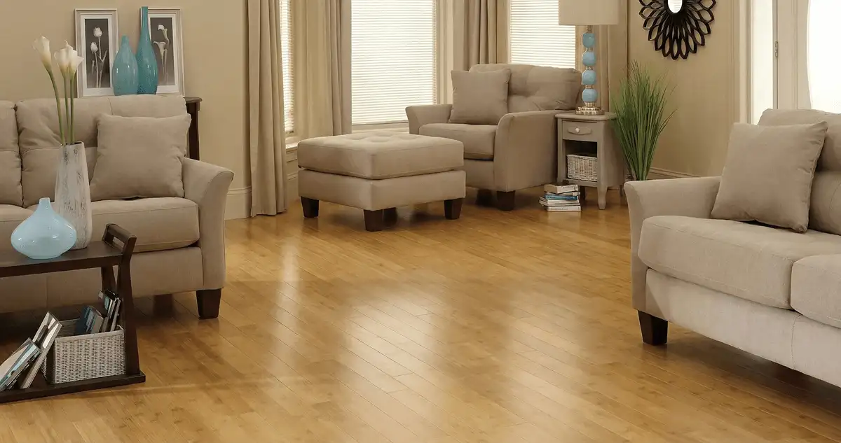 A living room with professionally installed bamboo flooring, beige sofas, a matching ottoman, a side table with a blue lamp, vases, framed pictures, and large windows with blinds and curtains in Gilbert, AZ
